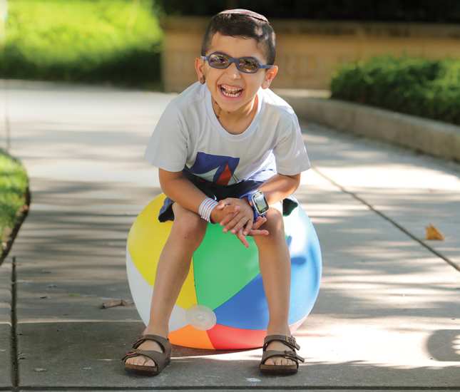 A young boy laughs and smiles while playing outside. He wears glasses and a kippa, and has a birthmark on his right ear and on the right side of his neck.
