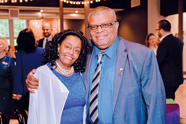 A woman and a man smile for the camera. The man is taller and to the right, and he has his right hand around the woman’s right shoulder. The woman is leaning into the man’s right shoulder. The man is wearing a blue suit, tie and glasses, and the woman is wearing a blue dress and pearl necklace.