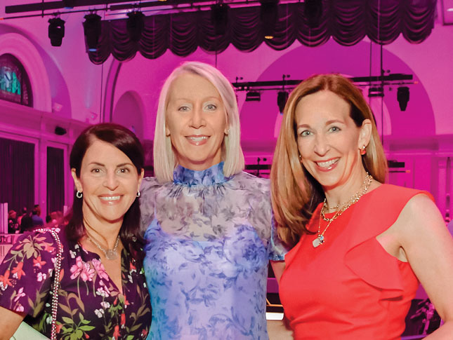 Photo of three smiling women at what appears to be a dressy event. On the left is a shorter woman in a dark purple floral dress. In the middle is a taller woman in a light purple floral dress. On the right is a medium-height woman in an orange dress.