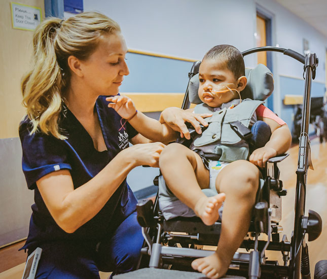 A nurse kneels to the side of a young boy and adjusts his seating in a mobility device.