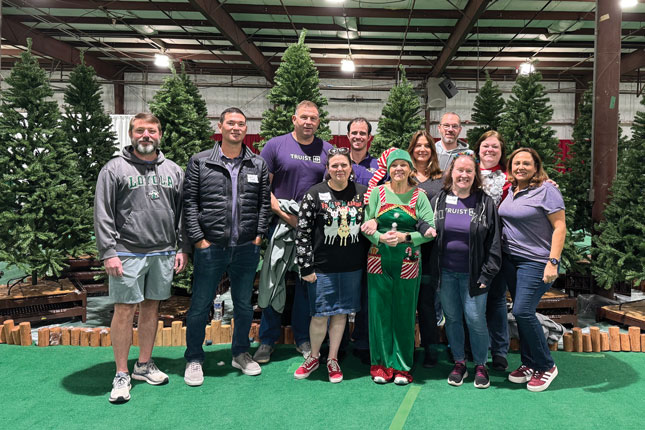 A group of 11 people of various ages, many wearing purple and one wearing a green Christmas elf costume, pose in front of a group of artificial trees inside a large indoor space, which looks like it might be a convention hall.