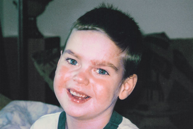 A young boy with dark blue-green eyes and blonde hair smiles for the photo. He is wearing a white T-shirt with dark green edging.