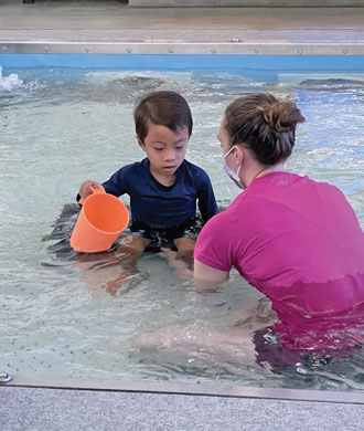 A little boy sits in a shallow pool holding a plastic orange pitcher. He appears to be engaged in water play. Assisting him is a woman wearing a face mask.