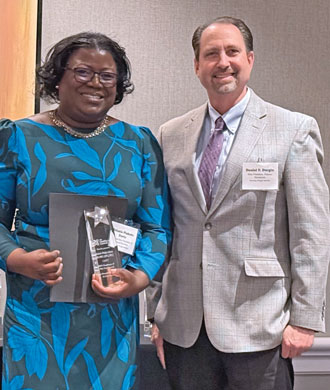 A smiling woman holds what appears to be an award. She wears a blue-and-gray dress. Next to her is a smiling man in a suit and tie.