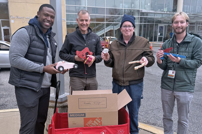 Four men, each holding a classic wooden toy, stand in front of a hospital building. In front of them is a red wagon with a box in it, presumably holding more wooden toys.