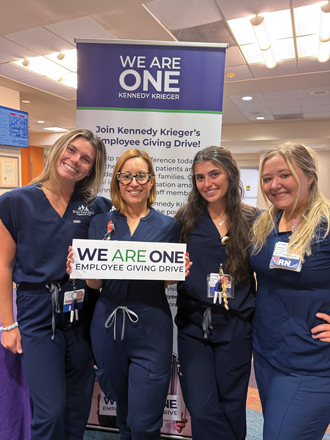Four Kennedy Krieger nurses smile while standing in front of a We Are One banner. One of them is holding a We Are One sign.