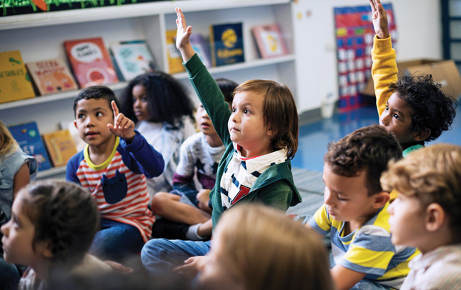 Three kids sitting on a floor in a classroom raise their hands. Several students sitting around them are not raising their hands.