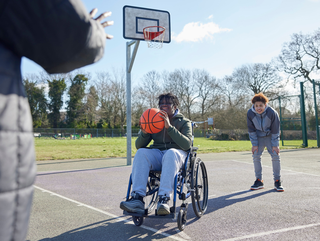 A teenager using a wheelchair plays basketball at an outdoor court with two other teenagers.