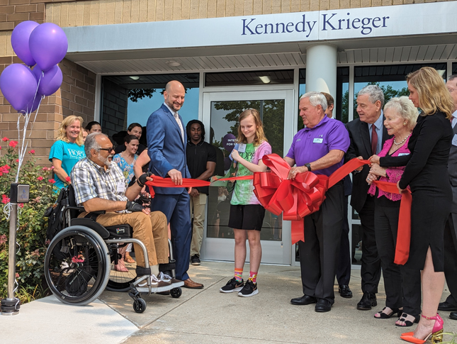 A group of at least 16 people of different ages gathers outside a glassed-in entrance with “Kennedy Krieger” above the doors. The group is holding a long, wide red ribbon with a giant bow in the center. A young person wearing a shirt that says “HOPE” is cutting the ribbon with giant scissors. Holding one end of the ribbon is a man sitting in a wheelchair. Some people are wearing suits, while others are wearing more casual clothing.