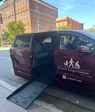 Un Toyota Sienna accesible para sillas de ruedas está estacionado en la acera con la rampa bajada. La camioneta adaptable es de color burdeos y tiene el logotipo de Kennedy Krieger Institute en la puerta del pasajero delantero.