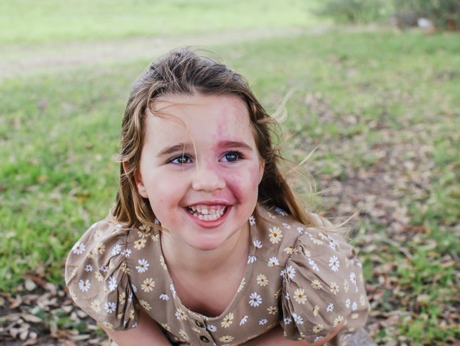 Rainey leans forward and smiles at the camera while sitting on the ground outside. 