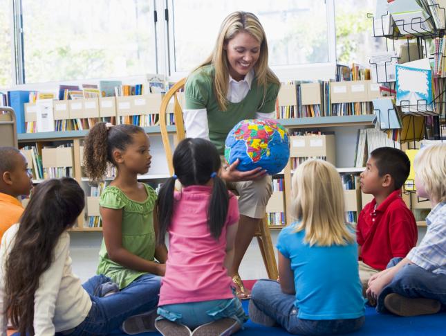 A photo taken in a classroom captures a teacher sitting in a chair and holding a globe and showing it to her students, sitting on the floor
