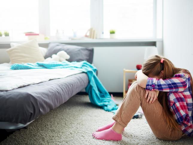 A photo of a girl sitting on the floor of her bedroom with her head down in her arms, in pain