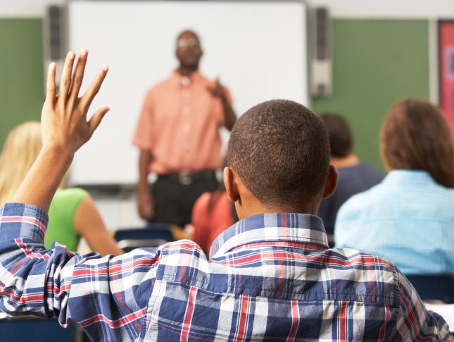 A photo of a student sitting in class, raising his hand and being called on by the teacher