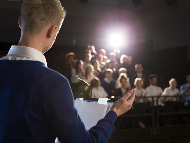 A photograph of a boy standing on a stage and presenting to an audience