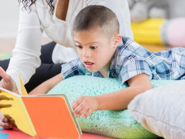 A child lays atop a pillow while reading a book, accompanied by an adult