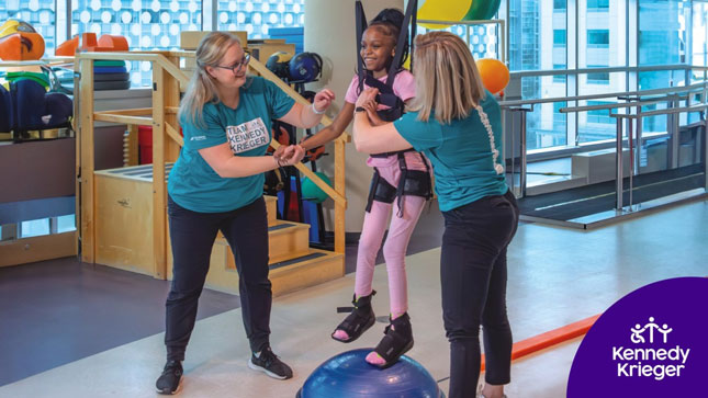 A girl smiles while doing physical therapy at Kennedy Krieger, with the help of two therapists.