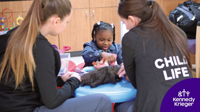 Two Child Life specialists sit at a table with a young girl. The girl is smiling as she plays with a doll.