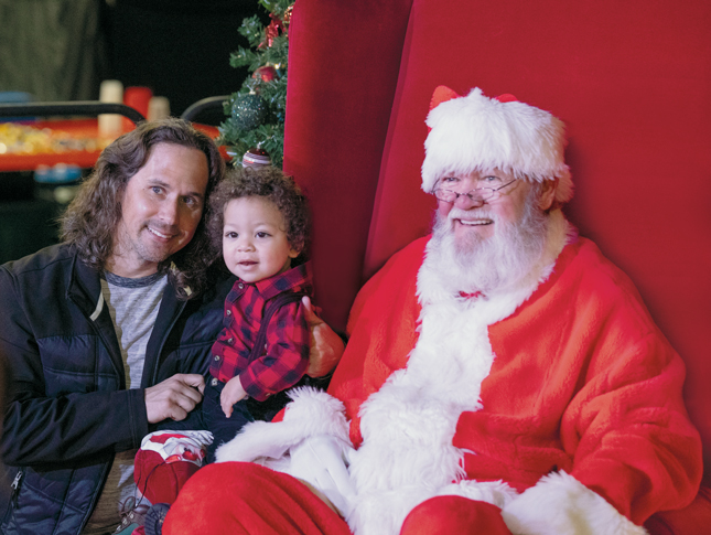 A father holds his toddler while kneeling next to Santa Claus at Festival of Trees.