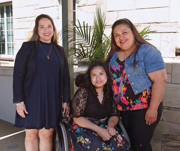 Una joven adolescente en silla de ruedas y vestida con un traje elegante, se encuentra entre dos mujeres. Ella se inclina hacia la mujer de la derecha, quien es su madre. Las tres sonríen.