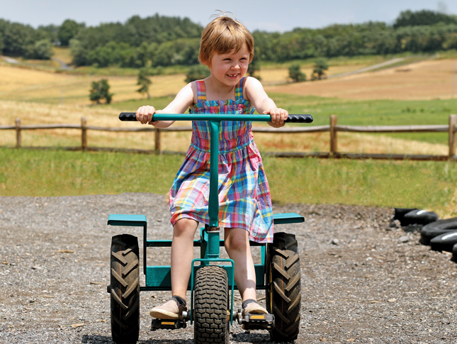 A smiling little girl with short blonde hair rides a tricycle in a rural setting.