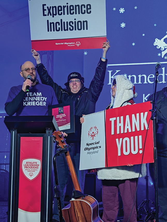 Kennedy Krieger President and CEO Dr. Bradley Schlaggar stands at a podium giving a speech while wearing a purple Team Kennedy Krieger Shirt. To his left is someone holding a Special Olympics Maryland sign that says Thank you! Behind Dr. Schlaggar is a man holding a Special Olympics Maryland sign that reads Experience Inclusion.