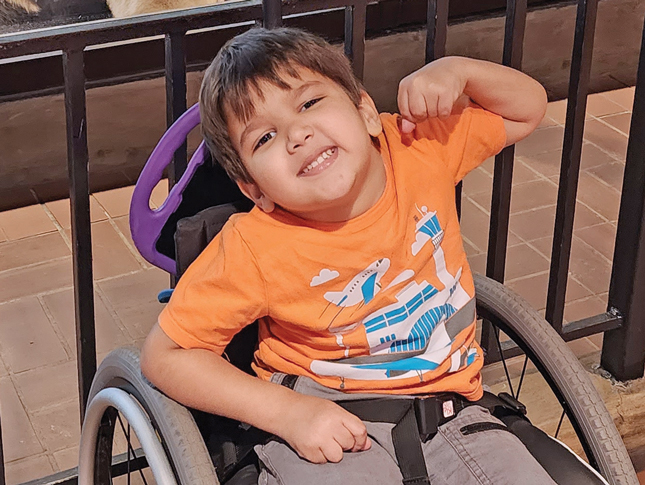 A young boy sitting in a wheelchair raises his left elbow while smiling. 