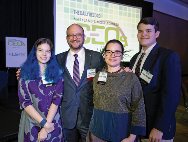Family photo of a teenaged girl and boy with their parents. Father and son are each wearing a suit and tie, and mother and daughter are each wearing a dress. Each family member is wearing a name badge. Behind them is a screen that says “The Daily Record—Maryland’s Most Admired CEOs.”