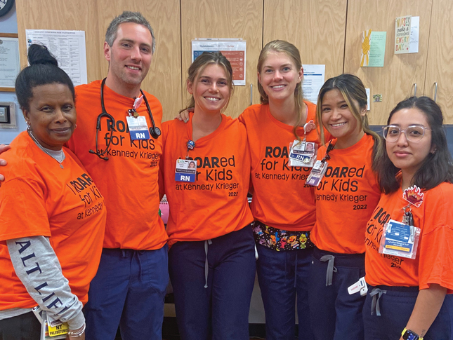 Six smiling people pose for the camera wearing bright orange shirts that say “I ROARed for Kids at Kennedy Krieger 2022.” Most of the people have badges on; some of the badges say “RN” on them. One of the people has a stethoscope around their neck.