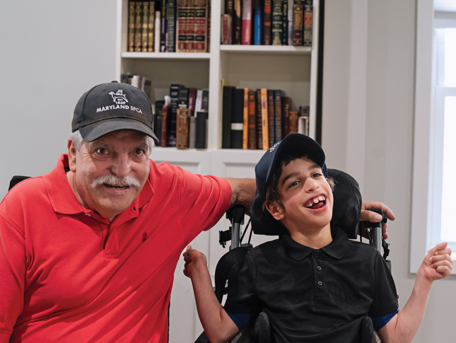 A man wearing a baseball hat that says “Maryland SPCA” on it sits beside a young boy who is also wearing a baseball cap. The boy is sitting in a wheelchair. The man’s left arm rests on the back of the wheelchair, behind the wheelchair’s headrest. Both the man and the boy are smiling. They are sitting indoors, in front of a bookshelf.