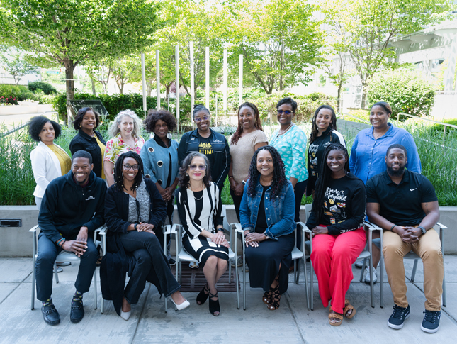 A group of 15 people pose in a garden-like setting. Six of them are sitting in chairs, while the other nine stand behind them. All are smiling, and most are people of color.