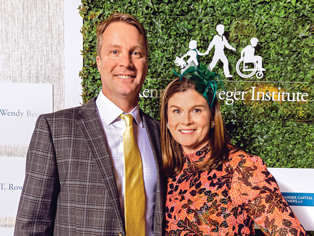 A husband and wife wearing fun formal wear pose for the camera, smiling. Behind them is a clear sign with Kennedy Krieger Institute’s logo on it. The sign hangs in front of a leaf-covered wall.