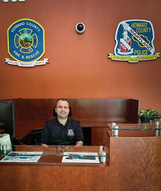 Andy sits behind the front desk of the Howard County Public Safety Training Center. Signs depicting the county's Fire & Rescue and Police badges are on the wall behind Andy.