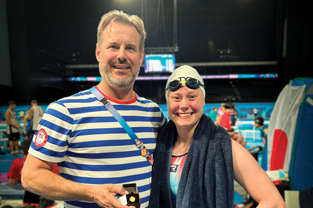 Taylor Winnett stands next to swim coach George Leatherman and smiles as the two stand poolside. She is wearing a swim cap and goggles.
