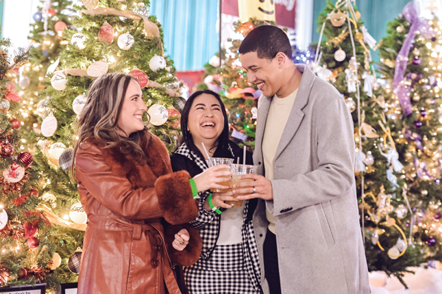 Three Tinsel on Tap attendees smile while standing in front of a grove of lighted trees.