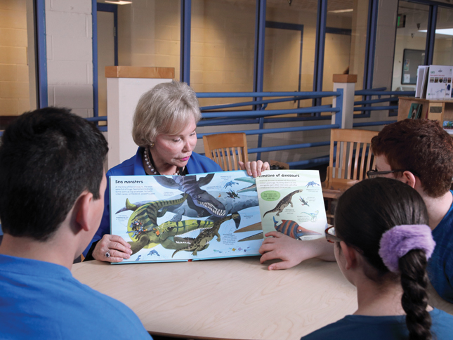 Nancy Grasmick sits at a table with students, holding out a book with illustrations of sea monsters and dinosaurs as she reads to them.