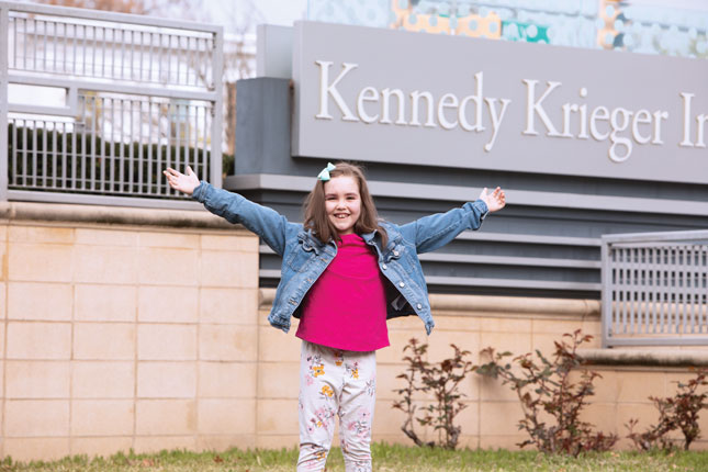 A young girl smiles with her arms outstretched. She has faint port wine-colored birthmarks across her forehead. A sign with the words “Kennedy Krieger” is in the background.