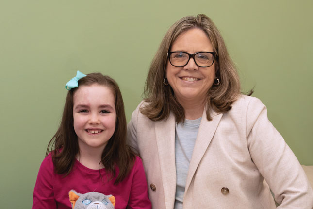 : A young girl with port wine-colored birthmarks across her face sits next to a woman in a suit. Both are smiling, and the girl is holding a stuffed animal.