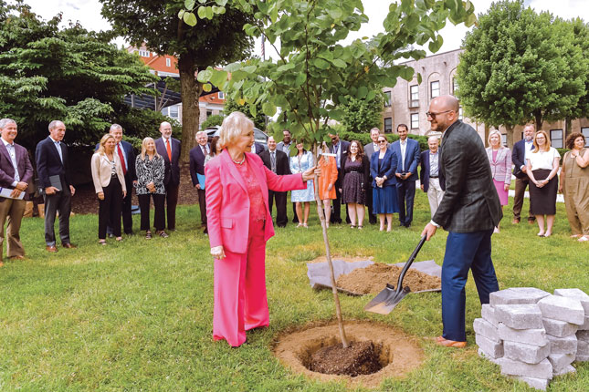 A woman and a man in business attire plant a tree, with a group of people standing behind them.