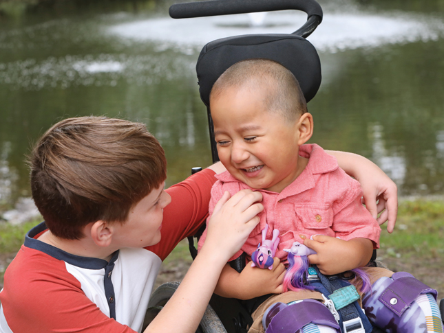 Two young boys, one a little older than the other, look at each other, giggling. The younger boy sits in a wheelchair.