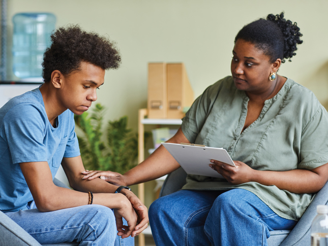 A woman holding a clipboard and with a concerned expression on her face places one of her hands on the arm of a teenage boy.