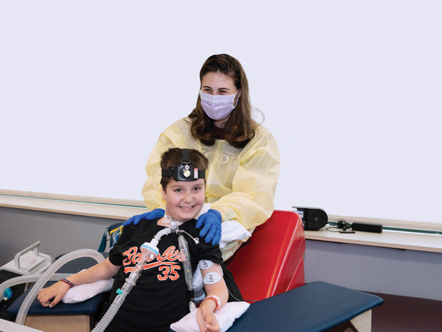 Posed photo of a smiling boy with a woman behind him. The woman is wearing a yellow medical gown and a face mask and has her hands on the boy’s shoulders. Her squinting eyes indicate she is smiling under her face mask. The boy is smiling and wearing a shirt that says “Orioles” on it. He has clear plastic tubes attached to his neck for breathing purposes and is wearing a head lamp-like device. Two electrodes marked “Leo” are attached to his left arm.