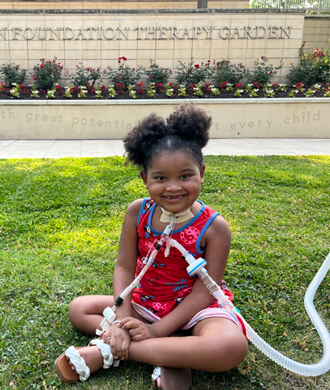 A young smiling girl sits cross-legged, with clear plastic tubes attached to her neck for breathing purposes.)