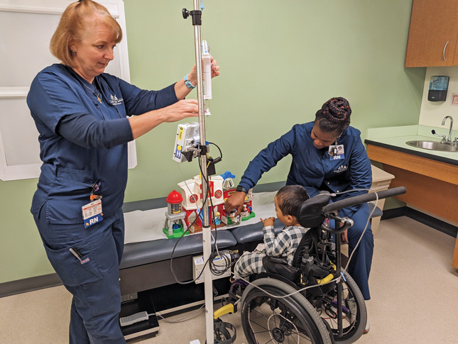 A small boy sits in a wheelchair in front of a table with a toy on it. To his right is a nurse helping him with the toy. The boy is hooked up to an IV infusion, which is being checked by a second nurse, to his left.