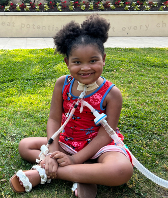 A young smiling girl sits cross-legged, with clear plastic tubes attached to her neck for breathing purposes.)