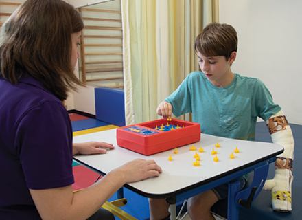 A photo of Brayden during a constraint-induced and bimanual therapy session at Kennedy Krieger with occupational therapist Lindsey Harris, in which his left arm is immobilized in a removable cast.