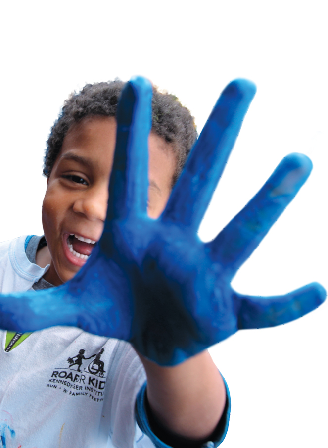 A child holds a paintbrush in his right hand while holding his left hand to the camera, with his palm covered in blue paint