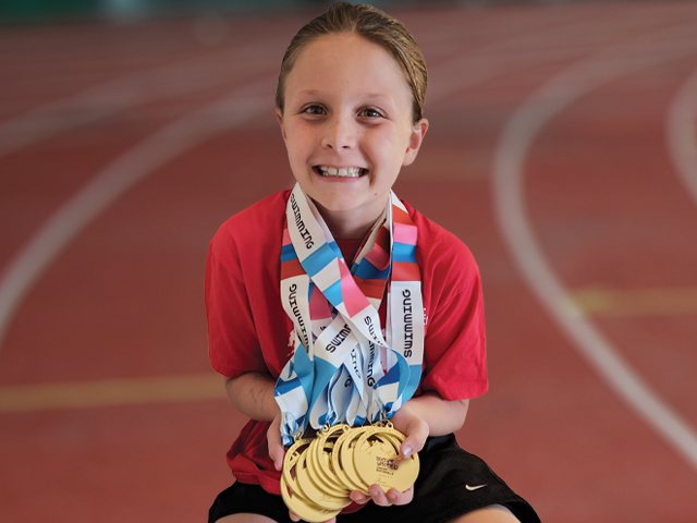 Penelope, smiling, wears nine medals around her neck, all of them gold, and with red, white and blue ribbons. Some of the ribbons can be seen to say “SWIMMING” on them.