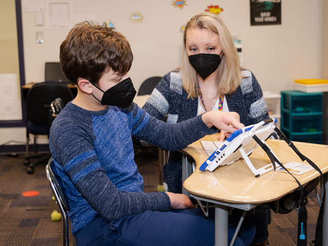 Jack sits at a student desk in a classroom with his teacher and uses a tablet-like communication device.”