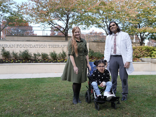 Violet sits in a wheelchair outside, with Dr. Ryan Gill holding the wheelchair to the left, and Dr. Mahim Jain holding the wheelchair to the right